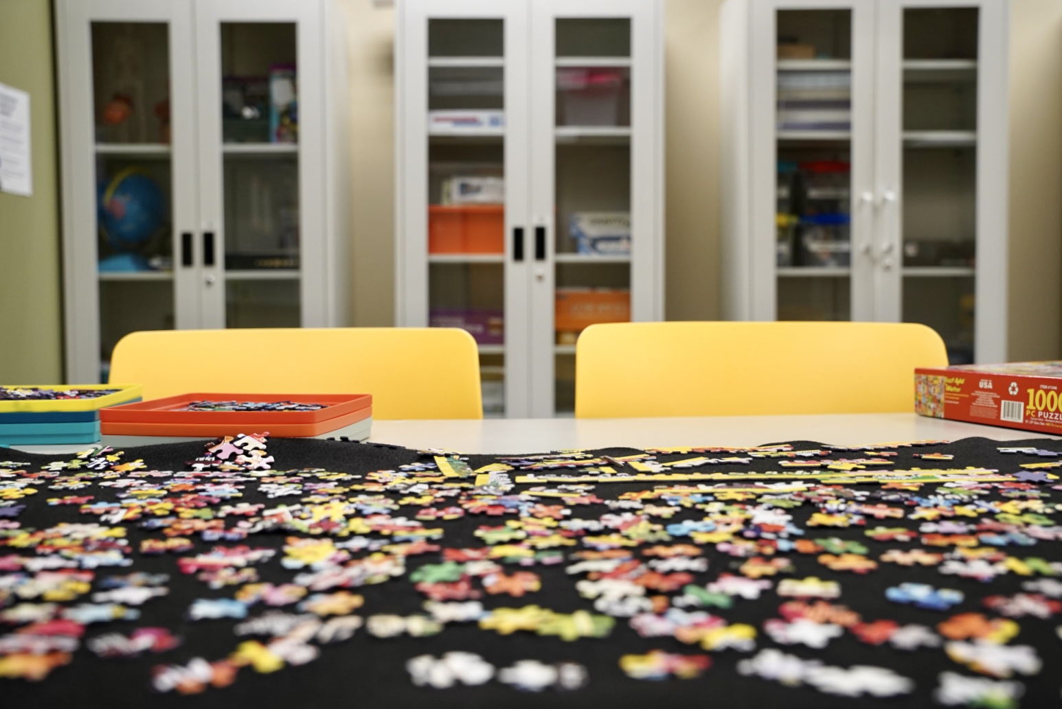 A puzzle is in the foreground and three bright glass-door cabinets filled with learning tools and games are in the background.